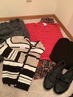 Six items including three tops (grey sweater, coral open-knit sweater, striped cardigan), a pair of black Mary Jane-style shoes, a black folded garment, and a colorful patterned fabric piece under the coral sweater.