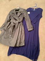 Full view of gray trench coat and blue dress on hangers displayed on floor