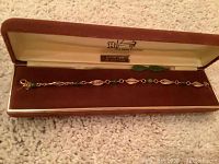 Bracelet displayed in open brown and cream box showing full bracelet length with mix of gold-filled links and jade beads plus green jade leaf charms.