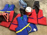 Photo showing 4 adult life vests in red and blue colors with yellow safety straps and a red dog life vest, along with a green sleeping bag visible in the background.