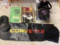 Image showing the Sony wireless headphones on charging base, two Chevrolet Corvette service manuals, a black Corvette-branded vinyl cover with yellow and red logo, black gloves, and a black Chevrolet cap on a concrete floor.