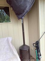 Full side view of upright propane patio heater with brown metal cylinder base under a grey protective cover, attached hose seen on the side.