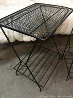 Photo showing black vintage mid century metal table with mesh top and slatted lower shelf on concrete surface.