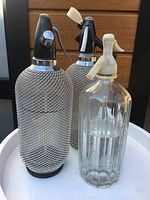 Three vintage glass seltzer bottles displayed together. Two have metal mesh covers and black siphon heads, one is clear glass with a white siphon head and etched design.