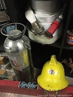 Photo showing silver metal fire extinguisher, coiled white fire hose with brass and red connectors, and yellow fireman helmet with emblem on custom shelf.