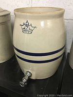 Photo shows the front side of the pottery crock with visible two blue stripes and spigot attached near the base.