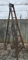 Full view of the vintage wooden A-frame ladder standing open outside against a wooden fence