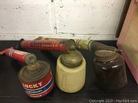 Photo of three vintage bug sprayers on black surface showing top and side angles; Lucky Continuous Sprayer, Hudson 2-Spray sprayer, and ceramic jar with metal lid.