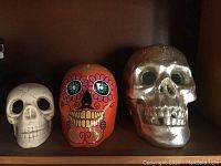 Three decorative skulls shown side by side on a wooden shelf: plain white skull, colorful Day of the Dead-style skull, metallic silver skull