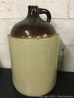 Full view of large vintage ceramic crock jug showing beige lower body and glazed brown upper neck and handle.
