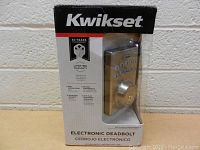Front view of the Kwikset electronic deadbolt lock in original box showing the lock and keypad through clear plastic.