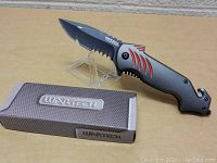 Wartech Shark folding knife displayed open on clear stand with box alongside on tan surface.