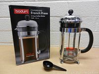 Overall view of Bodum Chambord French Press coffee maker with original packaging and coffee scoop on table against white brick wall.