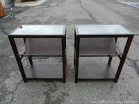 Front view of two matching rectangular end tables with glass tops and built-in angled wooden bookshelves, showing the dark wood frames and two shelves below the glass top.