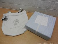 Folded blue damask queen sheet set packed and bound with labels on top, next to Threadmill Home Linen branded cream fabric tote bag with striped handles on table.