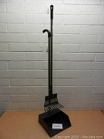 Full view of the metal dog pooper scooper with dustpan against a brick wall.