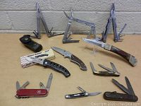 Lot of 9 pocket knives and 3 multi-tools arranged on a table, showing open tools and closed forms