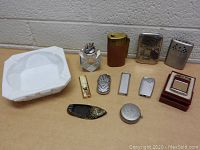Photo showing all 11 items including 9 vintage lighters and 2 ashtrays arrayed on wooden surface with gray wall background.
