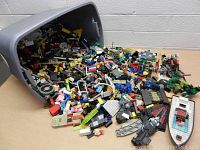 Large gray plastic tub tipped over with thousands of assorted loose Lego pieces spilling out onto a flat surface. Pieces include bricks, wheels, flat plates, and specialized elements in multiple colors.