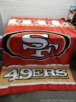 Large rectangular blanket with San Francisco 49ers logo and team name. Colors are red, gold, black, white. Blanket appears plush and new.
