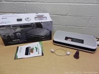 Photo showing NutriChef vacuum sealer device, original box, user manual, vacuum hose, and silicone bellow accessory on table.