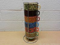 Set of six colorful ceramic coffee mugs stacked vertically in metal holder showing front design of each mug.