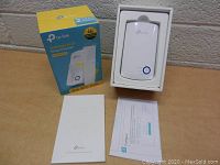 Photo showing the TP Link Wi-Fi range extender device inside its box, along with the user manual and printed setup guide, and the product box in the background.