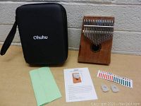 Overview of kalimba with black carry case, cleaning cloth, tuning stickers, finger protectors, and instruction manual on table surface next to a wall.