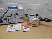 Photo showing MTA aluminum pressure cooker, original box, and instruction manuals on table with gray background.