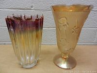 Two vintage glass vases side by side, one clear with purple and amber tones and vertical ridges, the other amber with embossed floral design and scalloped rim.