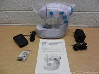 Top view of mini sewing machine with power adapter, foot pedal, extra accessories, and instruction manual all arranged on flat surface.