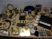 Wide view of a table with all miscellaneous collectibles including brass horn, metal cups, ceramic pots, pocket watches, toy gun, gaming set, and various decorative items.