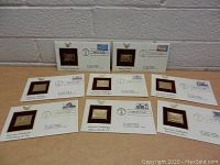 Lot of 8 First Day of Issue collectible envelopes with gold replica stamps mounted on burgundy background laid out on a table.