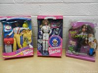 Three boxed dolls including Barbie Fire Fighter, Barbie Astronaut, and a Bratz Shrek doll on a tabletop against a white brick wall.