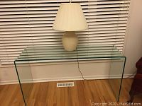 Straight-on view of clear glass console table against window blinds with lamp on top