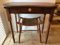 View of antique wooden table with single drawer featuring floral knob and matching woven seat stool beneath it.