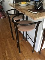 Front angle view of two bar stools under kitchen counter with brown cushioned seats and black metal frames.