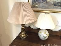 Two table lamps displayed side by side on a wooden surface, showing brass lamp with beige shade and ceramic floral lamp with white shade.