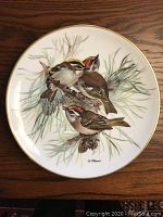 Collector plate featuring three birds with brown, yellow, and red markings perched on pine branches.