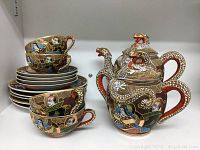 Full view of the tea service including four teacups stacked on six saucers with teapot, creamer, and sugar bowl showing the dragon motifs and colorful traditional scene paintings