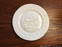 White ornamental ceramic plate with embossed fruits in center and ornate rim detail, lying flat on wood surface.