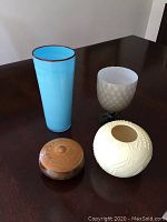 Four items displayed on a dark wood table: tall blue glass vase, white glass goblet-shaped vase, round white ceramic vase with textured pattern, small round wooden box with lid.