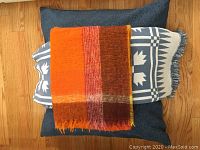 Blue floor cushion with two throw blankets stacked on top, one orange mohair and one blue and white patterned.