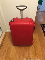 Red Samsonite hard shell suitcase upright with telescoping handle extended on wooden floor.