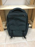 Full view of dark grey Samsonite backpack with front zippered pocket and top handle, standing on tile floor with wooden shelves behind