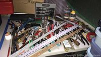 Overall view of assorted hand tools including screwdrivers, wrenches, rulers, pliers, and cutting tools arranged on a flat surface with measuring rulers placed on top.