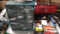 Photo showing green multi-drawer unit wrapped in plastic with screws and fasteners inside, red metal tool box, black tool box, and various items surrounding them.