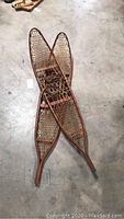 Photo of a pair of vintage wooden snowshoes lying crossed on a concrete floor, showing full length and detail of wood frame and rawhide webbing.
