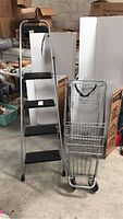 Overall view of 4-step stepladder next to foldable metal shopping cart in warehouse setting