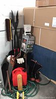 Photo showing multiple garden tools including post hole digger, pruning saw, clipper shears, gloves, boots, broom, hose, and gas can in storage area.